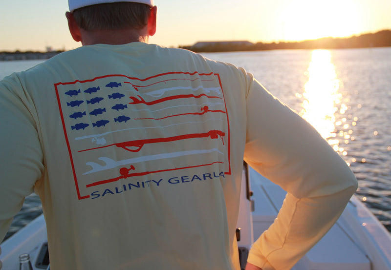 Salinity Gear performance SPF 50 sun protection dri-fit long sleeve fishing shirt. Pale Yellow shirt with screen printed Salinity Gear USA design. American Flag design created with spearguns surf boards and fish on the back. The front has The Salinity Gear logo with the American Flag inside of it.